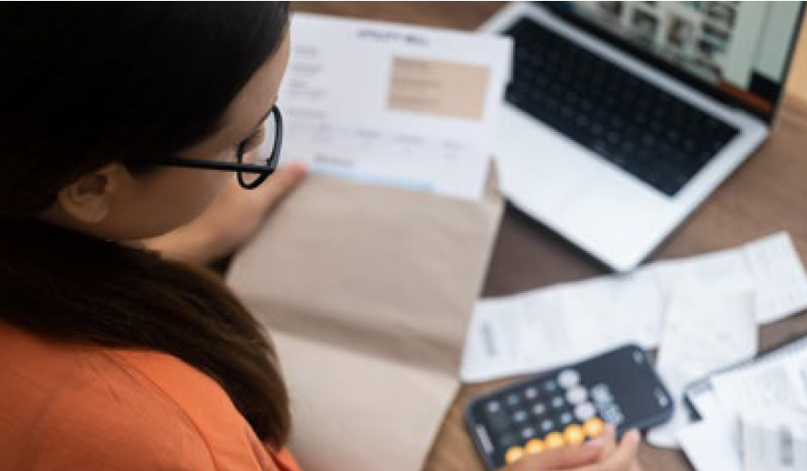 Person with glasses and orange shirt reading utility bill at desk with computer and calculator.