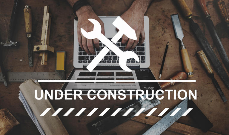 Under construction banner in foreground with person at laptop keyboard surrounded by work tools.